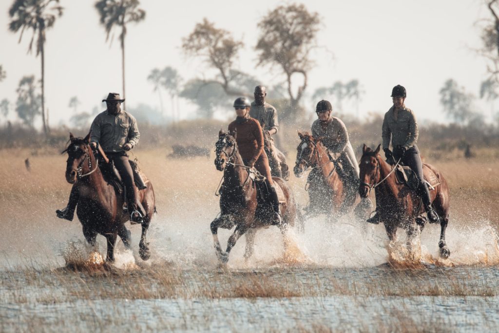 Eine Gruppe Reiter galoppieren durchs Wasser im Okavango Delta