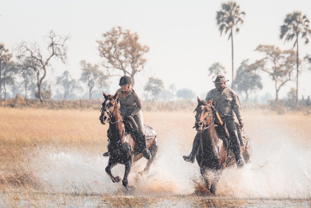 Frau und Ranger galoppieren im Okavango Delta durchs Wasser