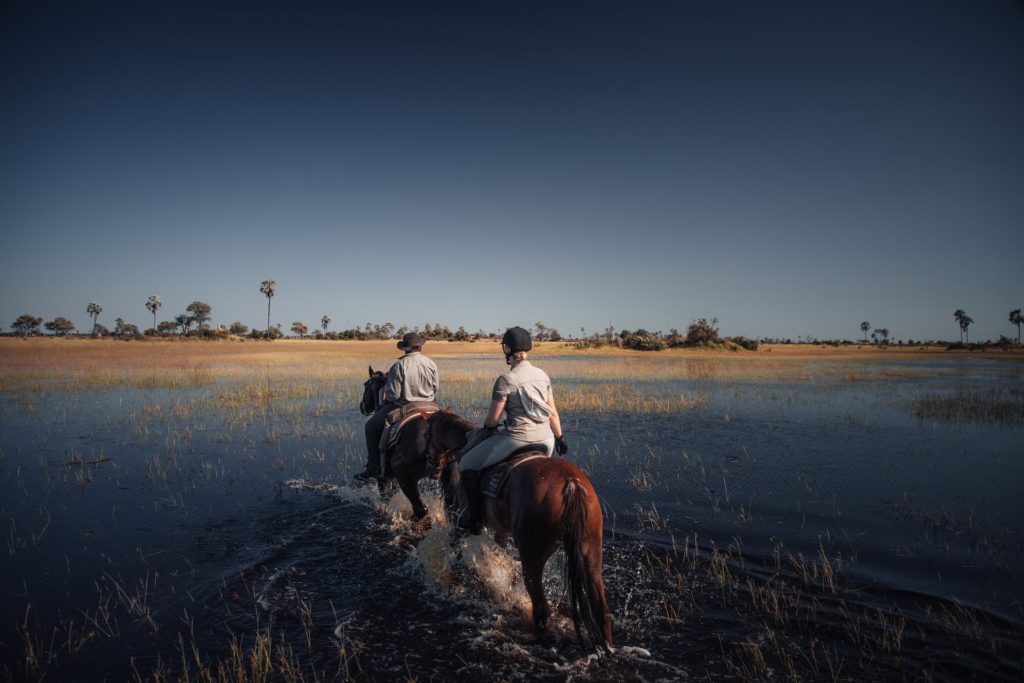 Frau reitet mit Ranger durchs Wasser im Delta Botswanas