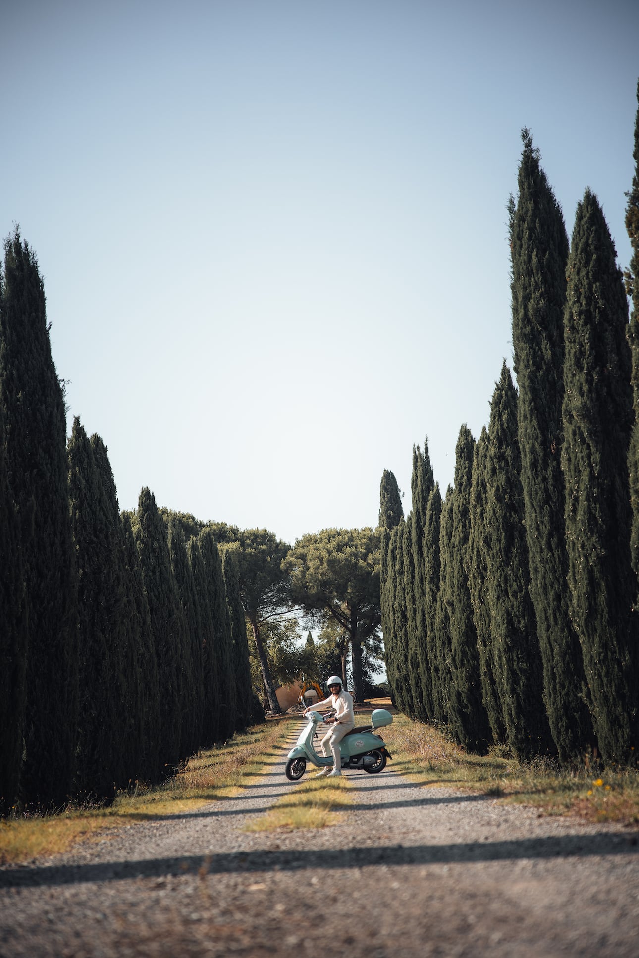 Mann fährt auf Vespa Roller durch Zypressen-Allee in der Toskana