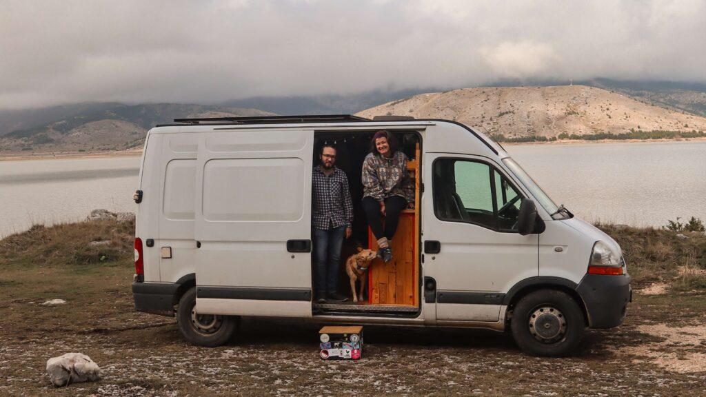 Mann und Frau sitzen in einem Van mit offner Tür vor einem See bei karger Landschaft
