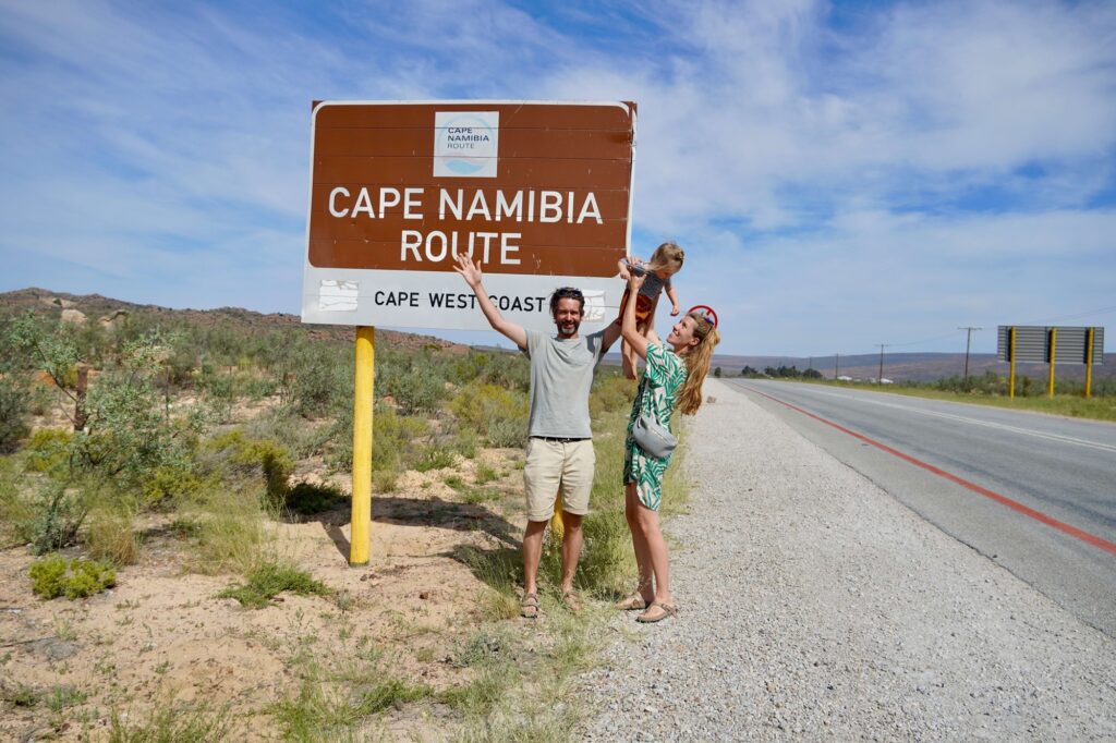 Frau und Mann mit Kleinkind am Straßenrand vor einem Verkehrschild mit der Aufschrift "Cape Namibia Route"